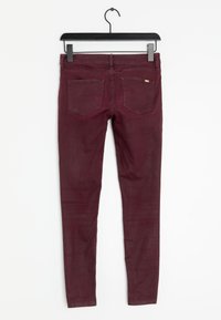 Jeans aderenti color ruggine con una texture liscia, design a cinque tasche e dettagli di cuciture sottili sulle tasche posteriori. Appesi a una gruccia nera.