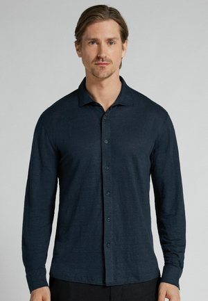 Mand med lyst brunt hår iført en marineblå langærmet skjorte med knapper foran mod en ensartet lys grå baggrund.