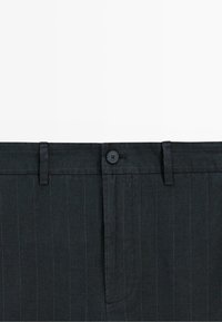 Zbliżenie na pas ciemnych spodni w prążki z zapięciem na guzik i szlufkami na pasek, pokazujące delikatne pionowe jasnoszare paski na tkaninie.