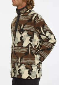 Pull en polaire dans des tons terreux. Présente un motif géométrique mixte, un col montant et une patte de boutonnage noire. Texture douce, coupe décontractée.