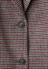 Tissu à carreaux gris et rouge avec une texture douce, présentant une fermeture à un bouton avec quatre trous. Design de col ajusté.
