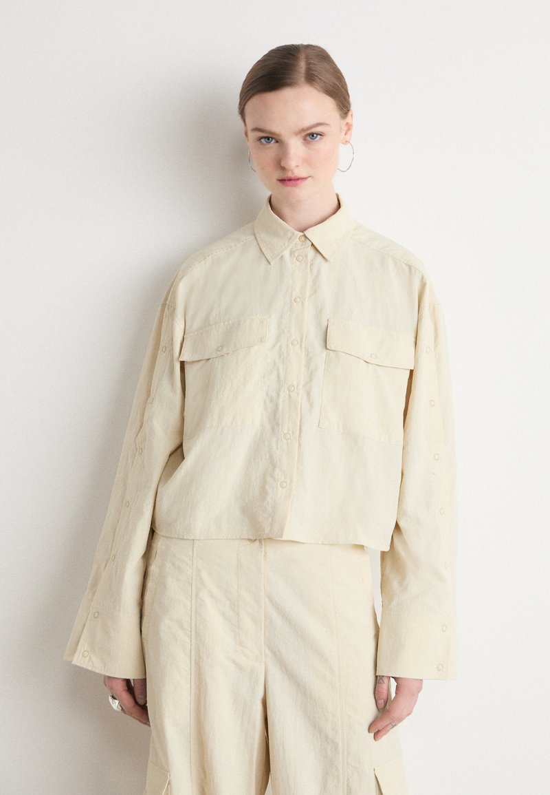 Beige, cropped button-up shirt with a pointed collar, two front pockets, and long sleeves with buttoned cuffs. Smooth, lightweight fabric.