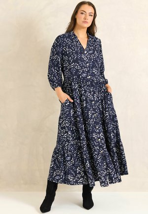 Femme portant une longue robe boutonnée bleu marine avec des motifs de feuilles blanches éparpillées et des bottes noires, debout les mains dans les poches.