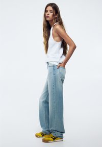 Vit ärmlös topp, ljusblå vida jeans och gula sneakers med svarta accenter. Slät tyg, lös passform, avslappnad stil.
