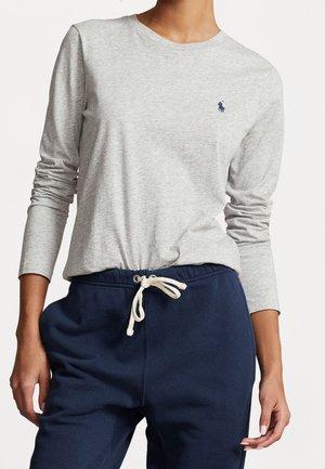 Personne portant un t-shirt à manches longues gris clair avec un petit logo bleu marine et un pantalon de survêtement bleu marine à cordon, debout avec les bras détendus.