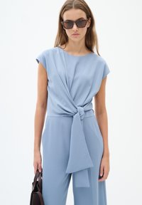 Tuta azzurra chiara con corpino aderente, maniche a cappuccio e dettaglio con fiocco sul davanti. Modella con una borsa a tracolla marrone scuro e occhiali da sole scuri.