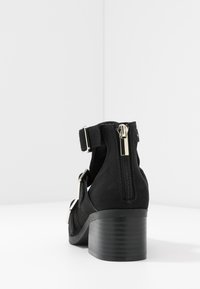 Stivaletto nero in finta suede con cerniera laterale, tacco chunky e dettaglio della cintura regolabile. Texture liscia e design semplice.