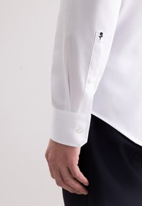 Camicia bianca con un motivo testurizzato, polsini abbottonati con un dettaglio di ricamo floreale nero decorativo e un design aderente.