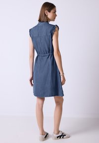 Denimmekko, jossa on lyhyet hihat, röyhelökoristeet, elastinen vyötärö, polven ylle ulottuva pituus ja vaaleansininen väri. Esitetty rentojen lenkkareiden kanssa.