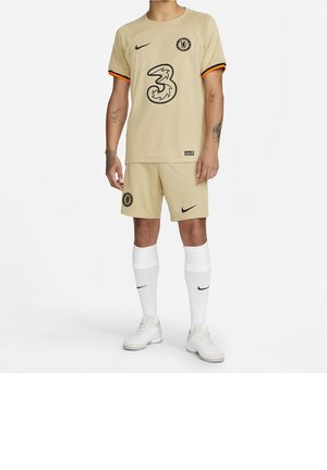 Personne portant un équipement de football Chelsea beige avec des logos Nike noirs, des chaussettes blanches et des baskets blanches, debout devant un fond blanc.