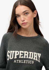 Szary sweter z dzianiny z okrągłym dekoltem i ściągaczami przy rękawach, z wyraźnym białym napisem "SUPERDRY ATHLETICS" na przodzie.