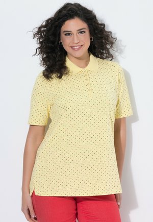 Uśmiechnięta kobieta z kręconymi włosami, ubrana w żółtą koszulkę polo w kropki z krótkim rękawem oraz czerwone spodnie na jednolitym tle.