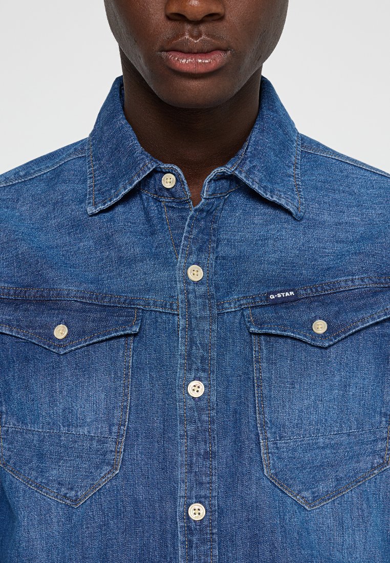 Chemise en denim bleu moyen avec un col classique, deux poches poitrine et des fermetures à boutons. Présente un tissu texturé et un détail de logo de la marque.