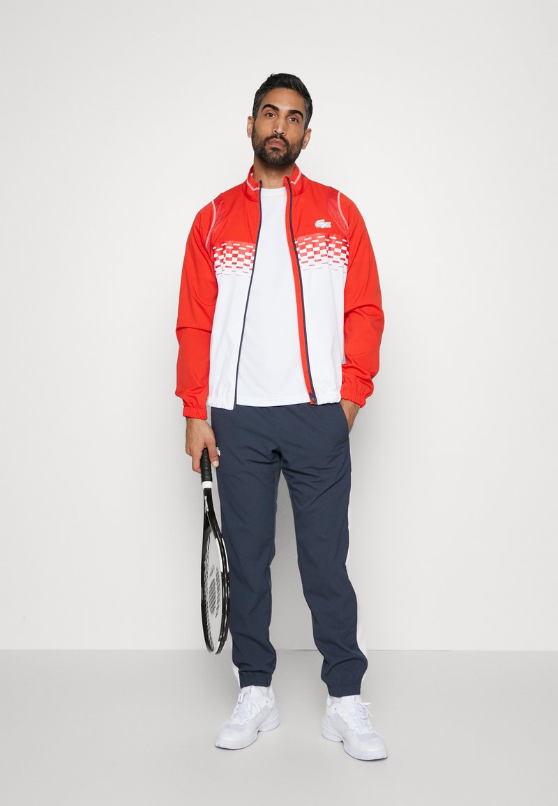 Giacca atletica rossa e bianca con design a quadri, abbinata a pantaloni blu e sneakers bianche, mentre tiene una racchetta da tennis.