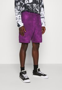 Shorts violet tie-dye avec deux poches avant et une poche patch sur la jambe. Associés à des baskets montantes noires et des chaussettes noires.