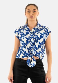Blusa bianca a maniche corte con un motivo floreale blu, dotata di un design con nodo frontale. Il tessuto sembra leggero e morbido.