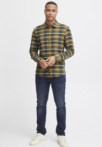 Flannel skjorte i grøn, gul og sort ternet mønster med knaplukning. Samtidig med mørke jeans og hvide sneakers. Glat tekstur.