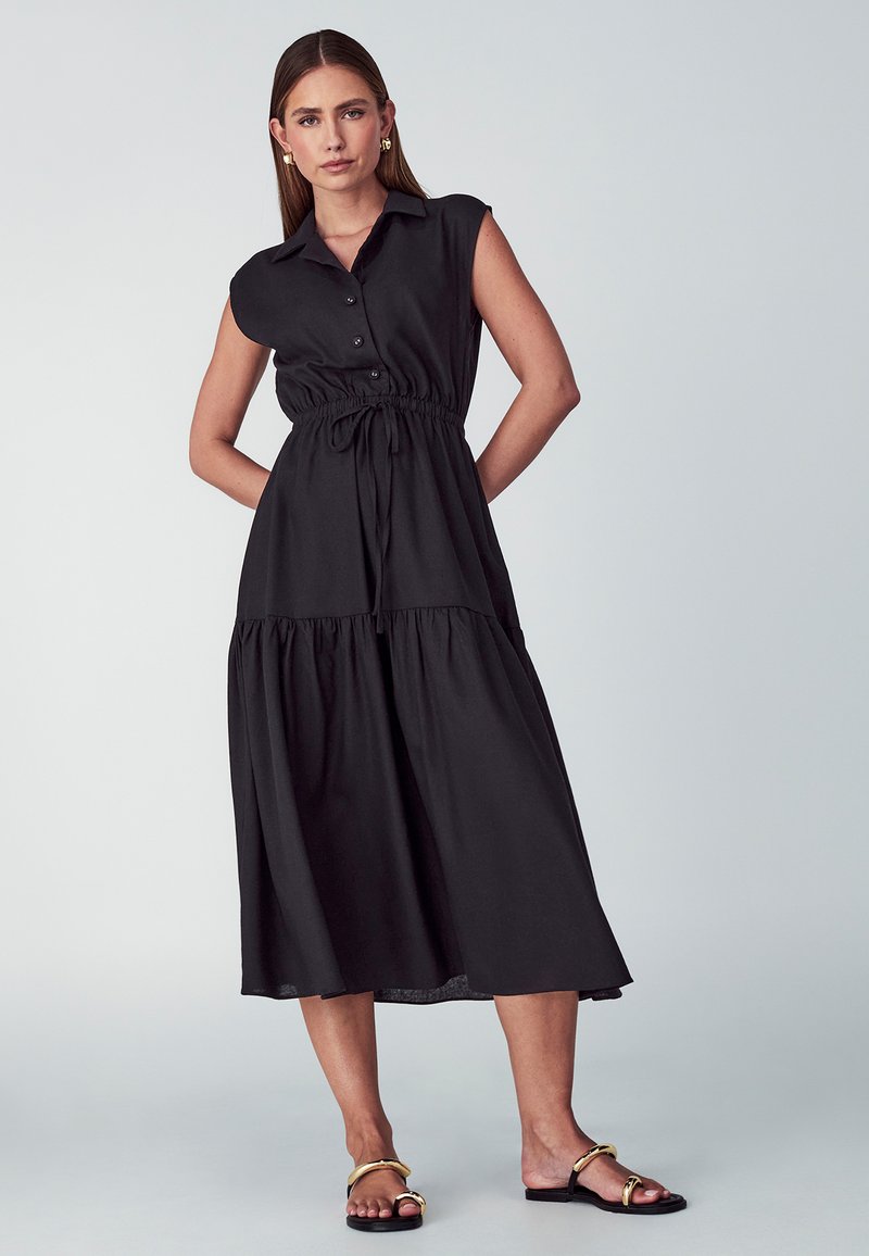 Czarna, bezrękawnikowa suknia z kołnierzykiem, guzikiem z przodu, elastycznym pasem i warstwowym dołem. Lekki materiał o gładkiej teksturze.