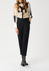 Blouse en satin avec motif géométrique en beige, crème et noir, dotée d'un foulard autour du cou ; associée à un pantalon noir sur mesure et des bottines à talons.