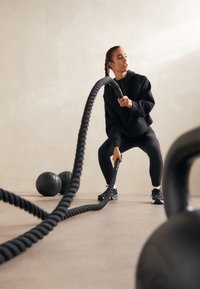 Czarna lina treningowa trzymana przez osobę w czarnej bluzie i legginsach, przygotowująca się do ćwiczeń, obok znajdują się czarne kettlebelle i piłki lekarskie.