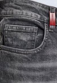 Los jeans de mezclilla grises presentan un bolsillo frontal, costuras visibles, una cintura negra y una etiqueta de marca roja. La textura es suave con un patrón desvanecido.