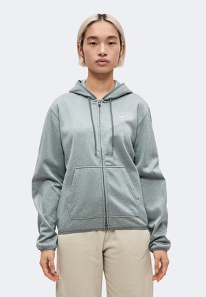 Modèle portant un sweat à capuche gris Nike zippé et un pantalon beige, debout devant un fond uni et clair.