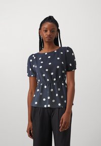 Blouse bleu marine à pois blancs, manches courtes bouffantes, taille froncée et encolure droite. Tissu lisse et léger.