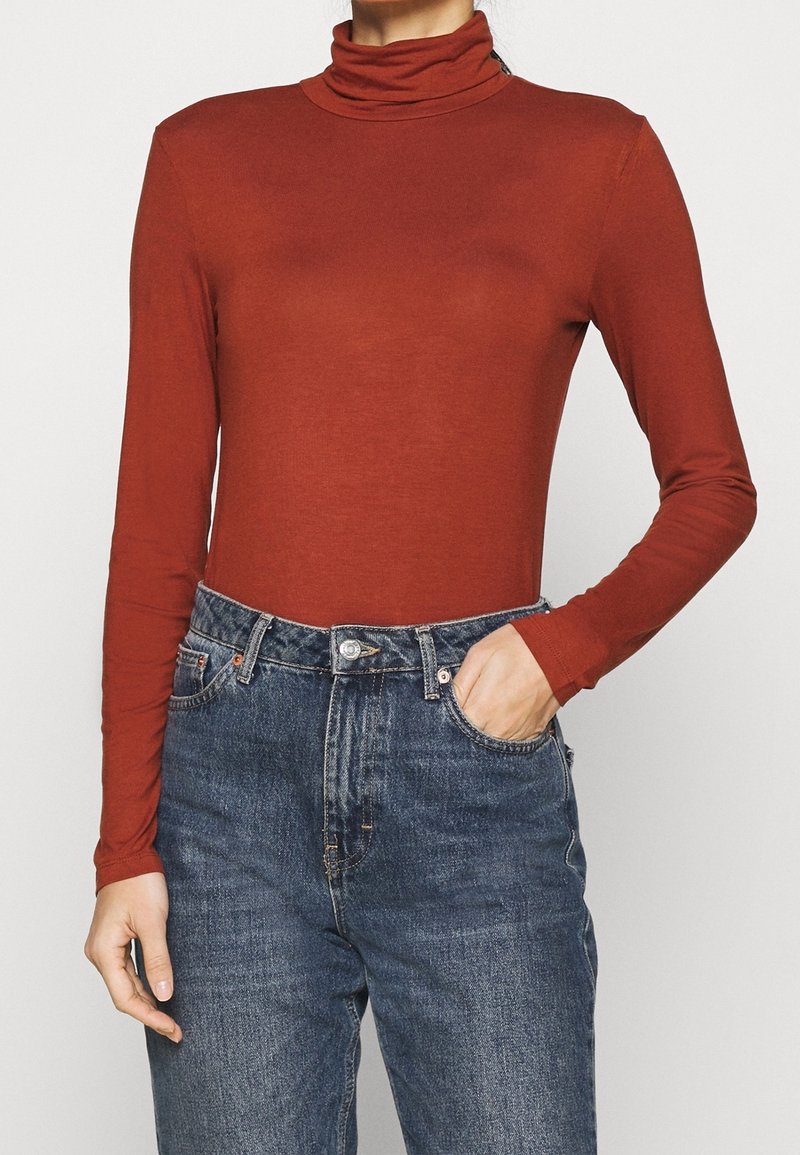 Osoba ubrana w rdzawego koloru golf z długim rękawem, wpuszczony w wysokie, niebieskie dżinsy, z jedną ręką w kieszeni.