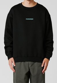 Persona vistiendo una sudadera negra con el texto "FORMER" en azul claro centrado en el pecho, combinada con pantalones verde oliva.