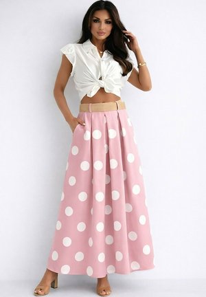 Femme portant un chemisier sans manches blanc noué et une jupe longue rose taille haute à gros pois blancs avec une ceinture beige, debout contre un mur blanc.