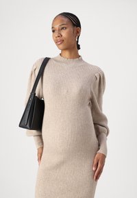 Beige ribbet strikket kjole med lange ermer og høy hals; brukt med en liten svart håndveske. Glatt tekstur, tettsittende silhuett.
