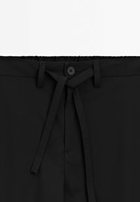 Schwarze Hosen mit elastischem Bund, vorderem Knopfverschluss und gebundenem Kordelzug. Glatte Textur, keine Muster oder sichtbare Hardware.