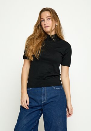 Frau mit langen, hellbraunen Haaren, die ein schwarzes Kurzarmshirt und eine lockere blaue Jeans trägt, steht vor einem einfarbigen hellen Hintergrund.