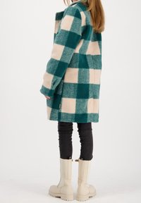 Cappotto a scacchi in teal e beige, realizzato in lana, con una vestibilità comoda, maniche lunghe e tasche laterali. Abbinato a jeans skinny neri e stivali beige.