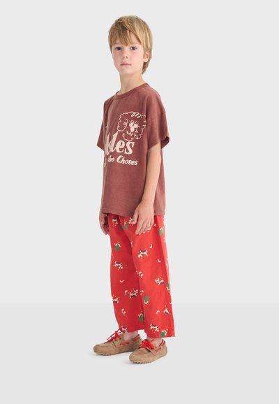 Ragazzo in piedi, che indossa una maglietta marrone con grafica, pantaloni rossi con motivi di mucche e cactus, e mocassini marrone chiaro con lacci rossi.