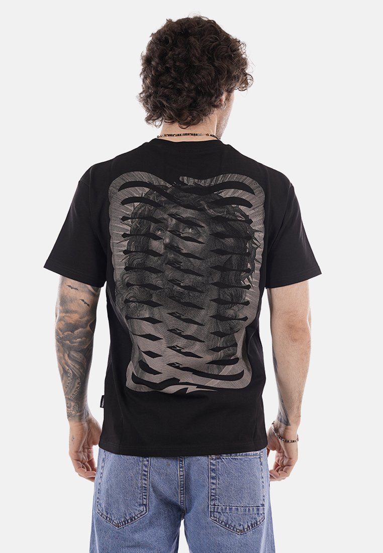 Uomo con capelli ricci che indossa una T-shirt nera con un design astratto di un volto stratificato sulla schiena, abbinata a jeans in denim azzurro.