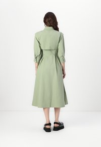 Rochie verde, de lungime medie, cu talie ajustată, manșete cu nasturi și o textură moale. Prezintă un spate plisat și un guler la gât.