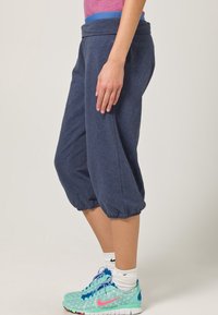 Tmavě modré capri kalhoty s elastickým pasem, manžetami na nohavicích a měkkým materiálem. Nošené s tyrkysovými a růžovými sportovními botami.