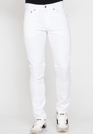 Jeans bianchi slim fit indossati da una persona abbinati a sneakers bianche e beige, in piedi davanti a uno sfondo chiaro e uniforme.