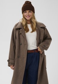 Brązowy płaszcz z mieszanki wełny z dużymi klapami, podwójnymi guzikami i bocznymi kieszeniami, noszony na jasnym swetrze i niebieskich spodniach z wysokim stanem, do tego brązowa dziana czapka.