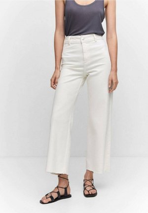 Žena v bílé širokých kalhotách s vysokým pasem, tmavě šedém tílku a černých sandálech s pásky na nízkém podpatku na jednobarevném bílém pozadí.