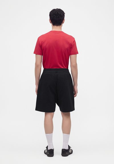 Homme de dos portant une chemise rouge à manches courtes, un short noir aux genoux, des chaussettes blanches et des baskets noires sur un fond uni.