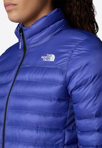 Giacca isolante blu con sezioni trapuntate, chiusura con zip, colletto alto e logo sul petto. Il materiale appare leggero e con una texture.