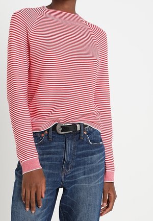 Osoba ubrana w czerwono-białą pasiastą bluzkę z długim rękawem, wpuszczoną w niebieskie jeansy z wysokim stanem i czarnym paskiem.