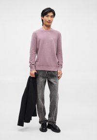 Lys pink strikket sweater med ribbede manchetter og kant, parret med grå denim bukser. Sort blazer holdt i venstre hånd, sorte sko fuldender outfittet.