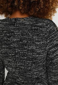 Femme aux cheveux bouclés portant une veste en tweed noir et blanc texturée, vue de dos et des épaules.