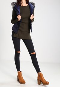 Marinkappad väst med pälsfodrad huva över en olivgrön ribbad tröja, svarta trasiga skinny jeans och bruna klackstövlar.