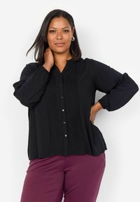 Blusa nera con scollatura a V, chiusura con bottoni e pieghe verticali nella parte anteriore, dotata di maniche lunghe con polsini elasticizzati.