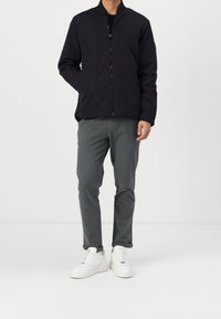 Uomo che indossa una giacca nera con zip, pantaloni grigi con risvolti e sneakers bianche, in piedi contro uno sfondo bianco uniforme.