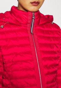 Rote Pufferjacke mit hohem Kragen, ausgestattet mit gestepptem Textil, Reißverschluss und dekorativen Zugbändern an der Kapuze. Silberne Hardware-Akzente.